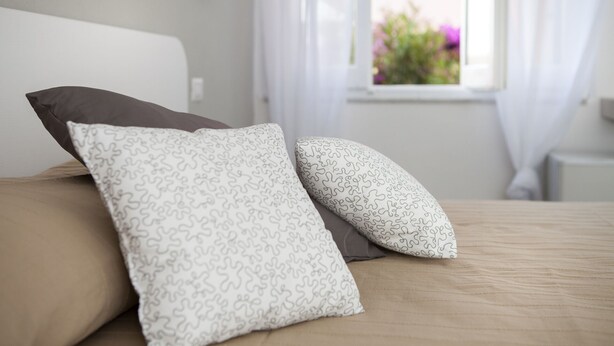 A bed with two pillows, a window with curtains, and a view of flowers outside.