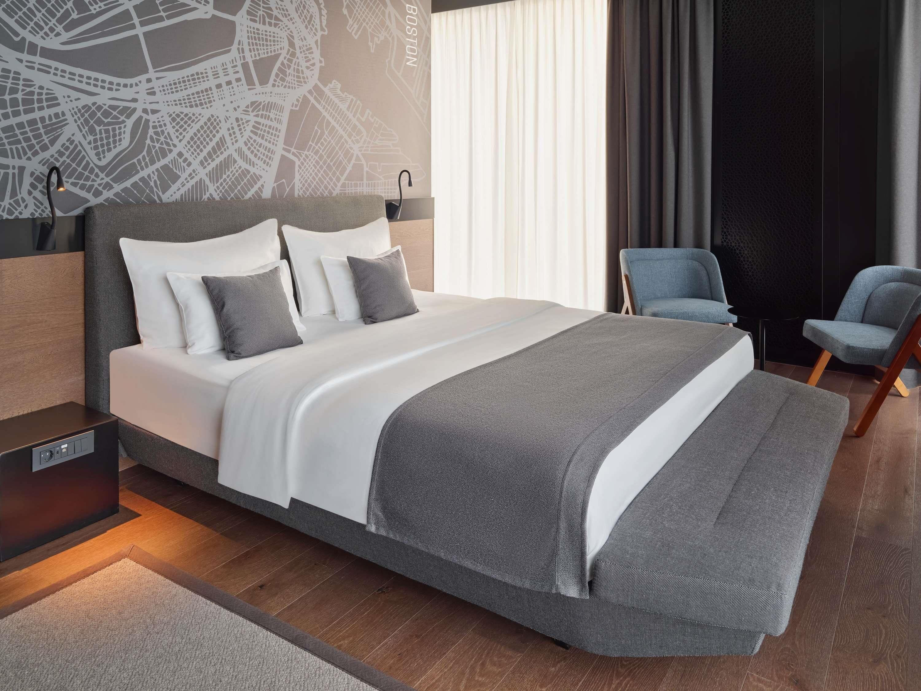 A modern hotel room with a large bed, a cityscape wall, a desk with a lamp, and a chair.