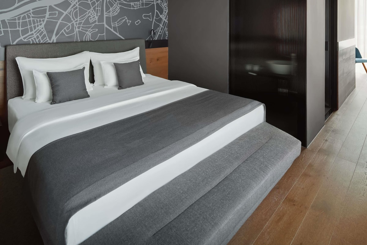 Een moderne hotelkamer met een groot bed, stadsgezicht kunst aan de muur, een donker schuifdeur en een houten vloer.