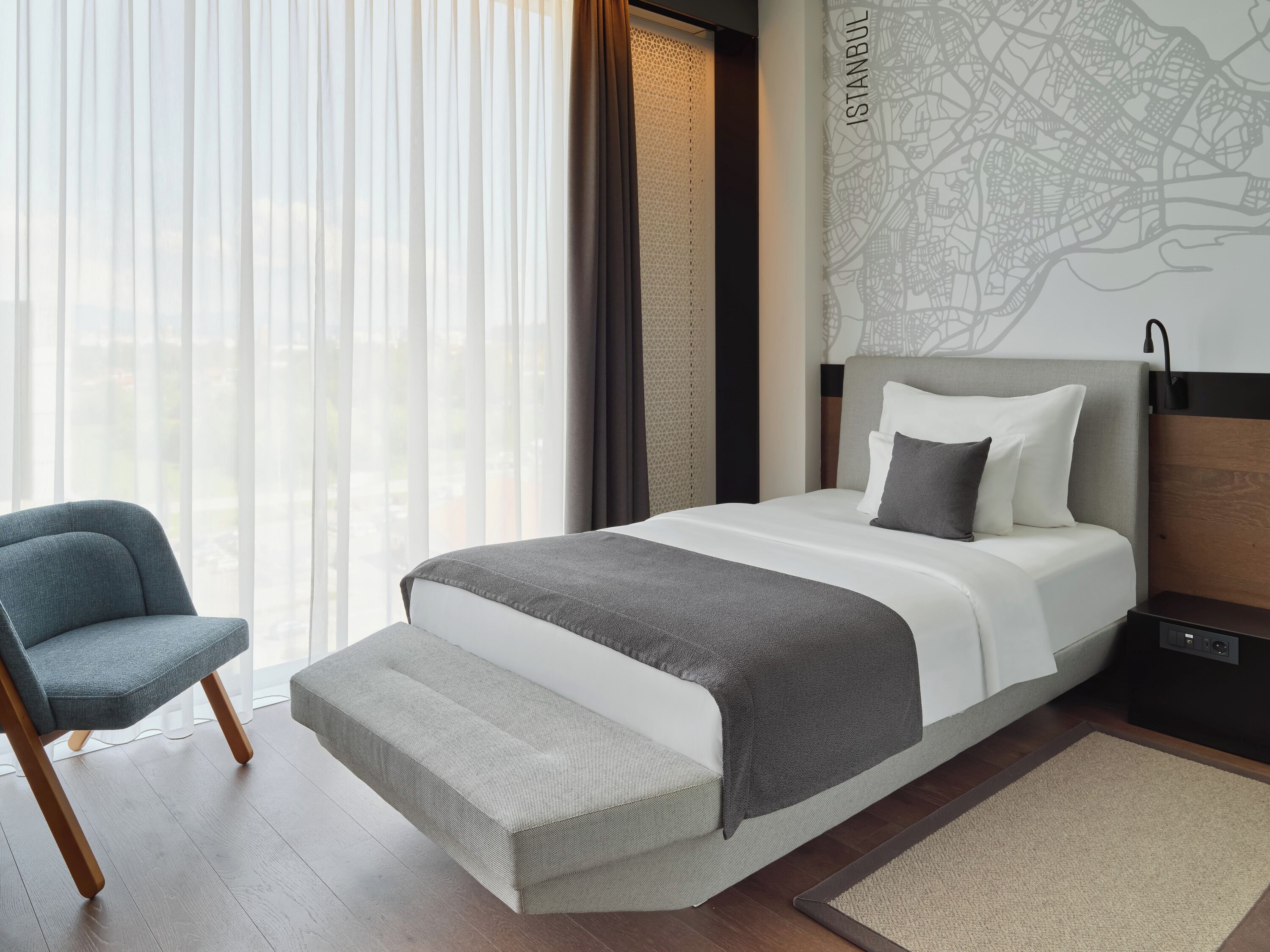 Una habitación de hotel moderna con una cama grande, una silla, un mapa en la pared y vistas a la ciudad.