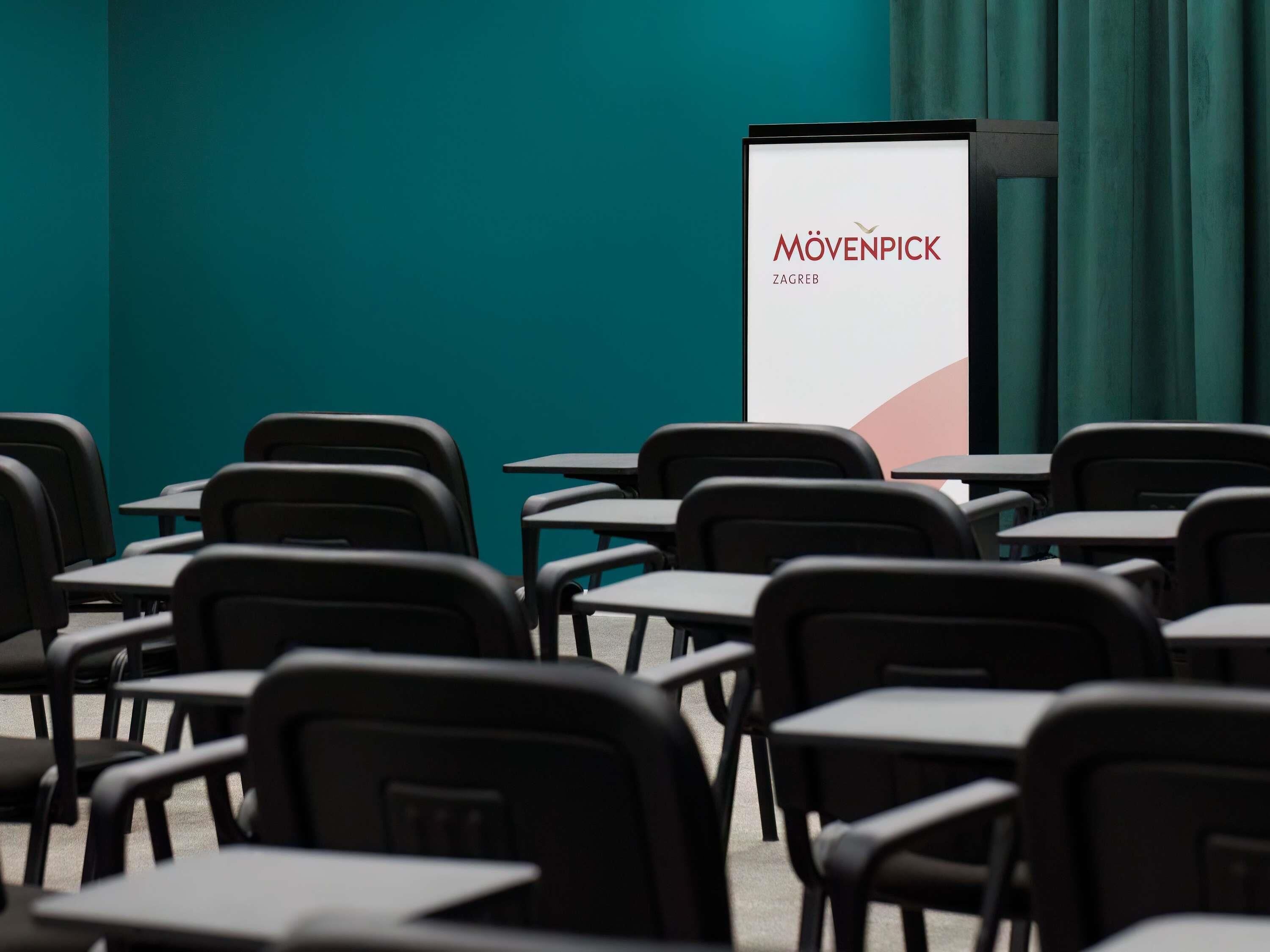 Una sala de conferencias con filas de mesas y sillas, una pantalla para presentaciones y una pared color turquesa.