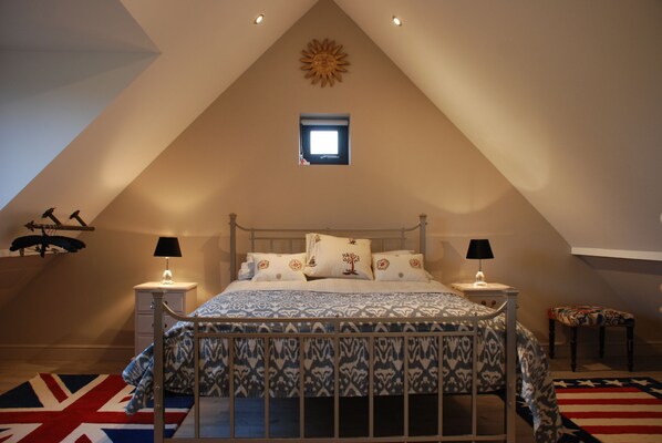 A bedroom with a sloped ceiling, a bed with patterned bedding, two bedside lamps, a small window, and a decorative sun on the wall. - Spring Cottage (Salisbury)