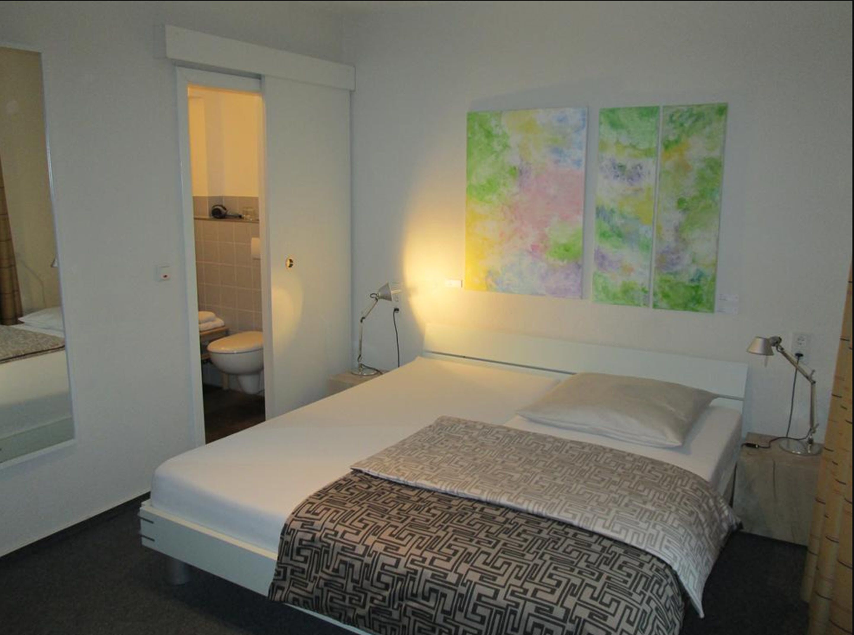 Ein Schlafzimmer mit Bett, Nachttischlampe, einem durchgehenden Türspalt sichtbaren Badezimmer und abstrakter Wandkunst.