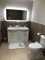 Um banheiro moderno com pia branca, espelho e vaso sanitário. Há uma toalha sobre um tapete branco, e uma lixeira é visível.