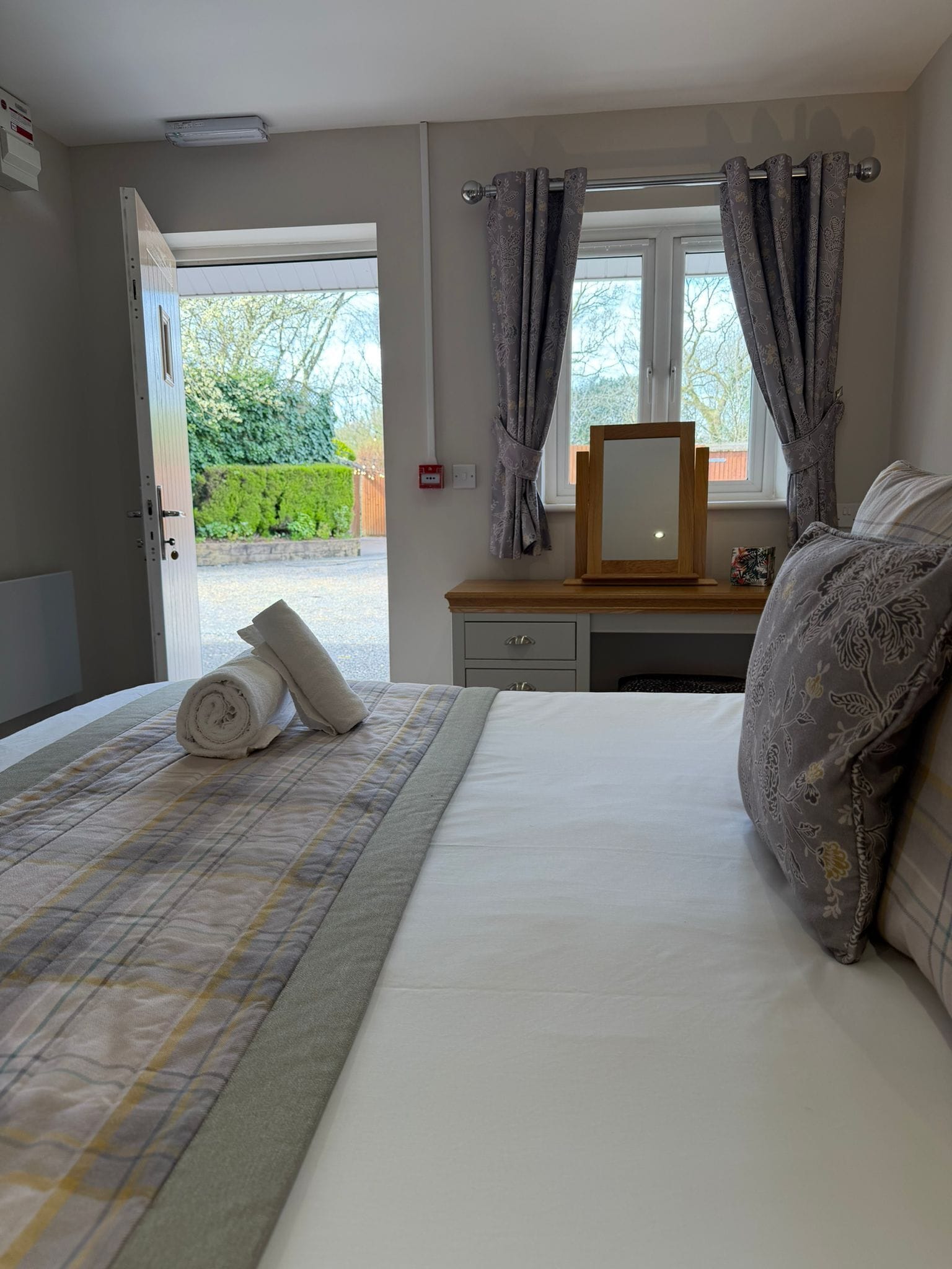 A hotel room with a bed, a bedside table with a mirror, a window with curtains, and a view of the outdoors.