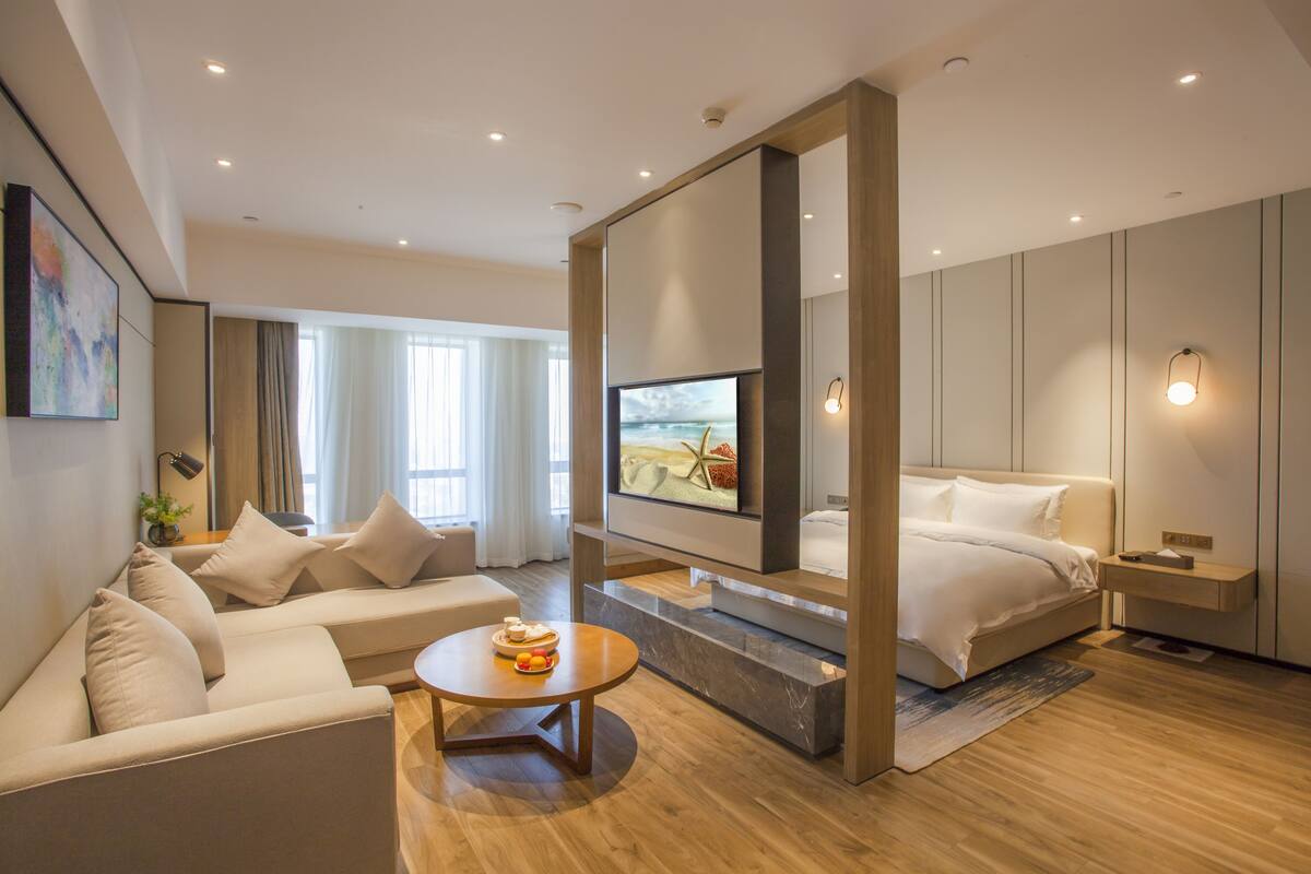 Une chambre d’hôtel moderne avec un canapé, une table basse ronde, un lit et un téléviseur fixé au mur.