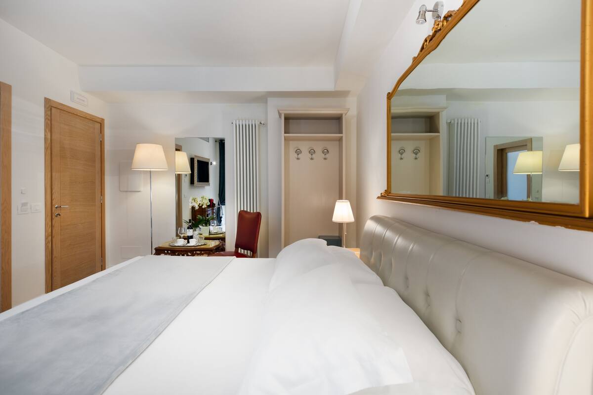A hotel room with a large bed, a wooden door, a mirror, a chair, and a desk with a lamp.