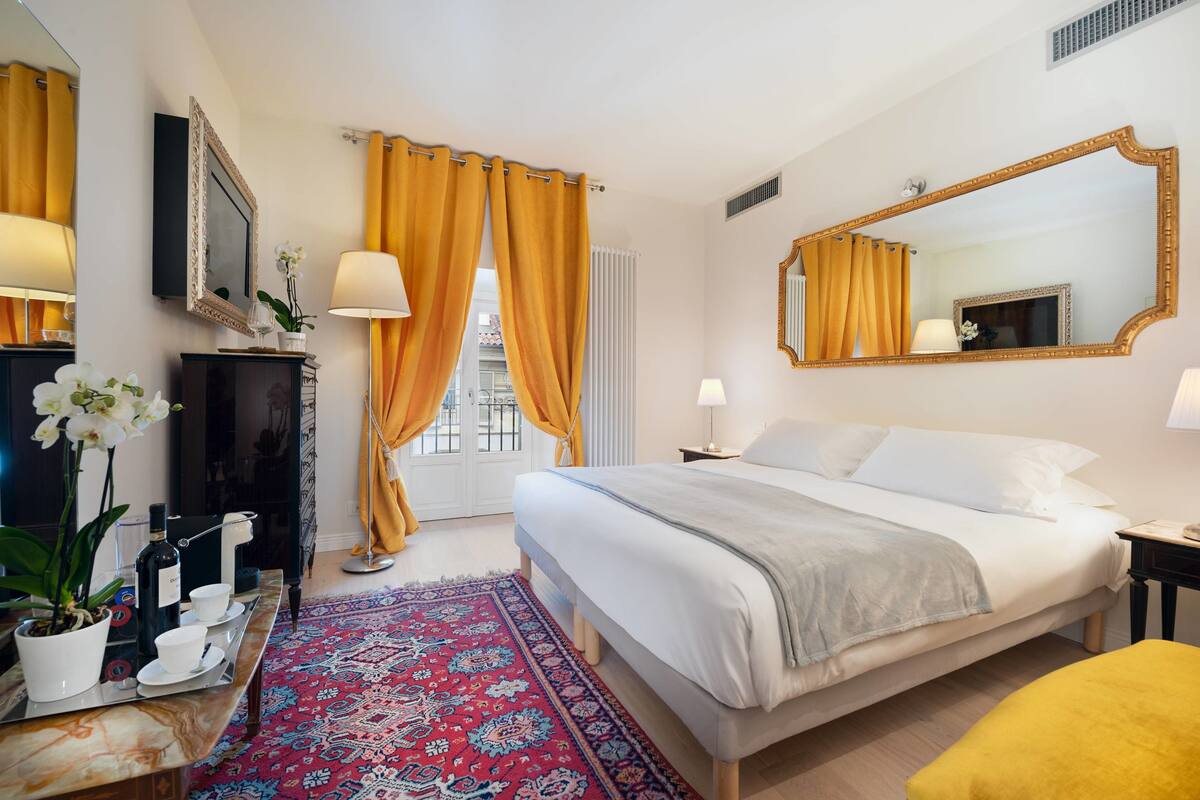 A hotel room with a bed, a TV, a mirror, a window with curtains, a small table with a lamp, and a potted plant.