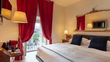 A hotel room with a large bed, red curtains, a window with a view, a bedside table with a lamp, and a mirror above the bed.