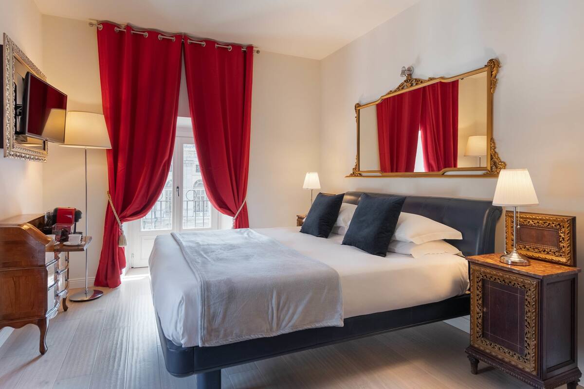 A hotel room with a large bed, red curtains, a mirror, and a bedside table with a lamp.