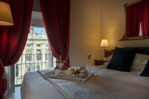 A hotel room with a bed, a tray with breakfast items, a window with red curtains, and a view of a building outside.