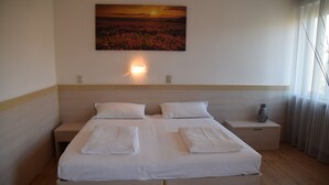 Een tweepersoonsbed met witte lakens, twee nachtkastjes, een wandlamp en een schilderij van een bloemenlandschap bij zonsondergang.