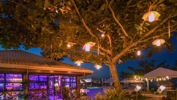 Une vue nocturne d’un complexe hôtelier doté d’une piscine, illuminée par un éclairage d’ambiance et entourée d’une végétation luxuriante.