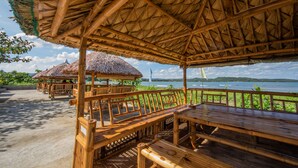 Un espace extérieur aménagé avec des sièges en bois et des parasols en chaume, offrant une vue sur une étendue d’eau et une île lointaine.