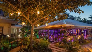 Un espace extérieur bien éclairé, avec un pont en bois, un sentier et un bâtiment doté d’une façade en verre et d’un éclairage violet.