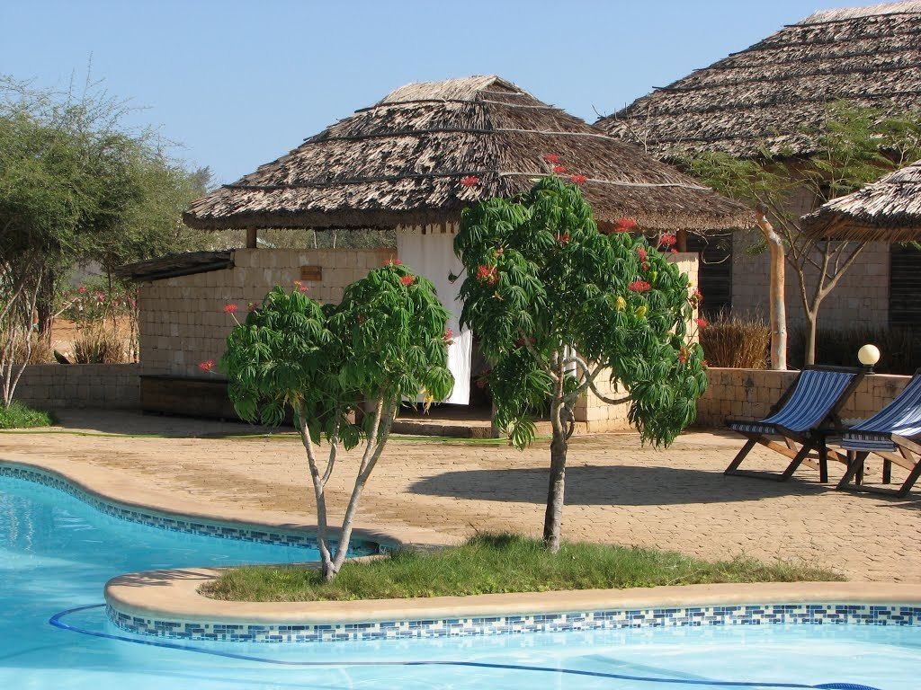 A swimming pool with a tiled edge, surrounded by a sandy area with lounge chairs and thatched-roof structures.