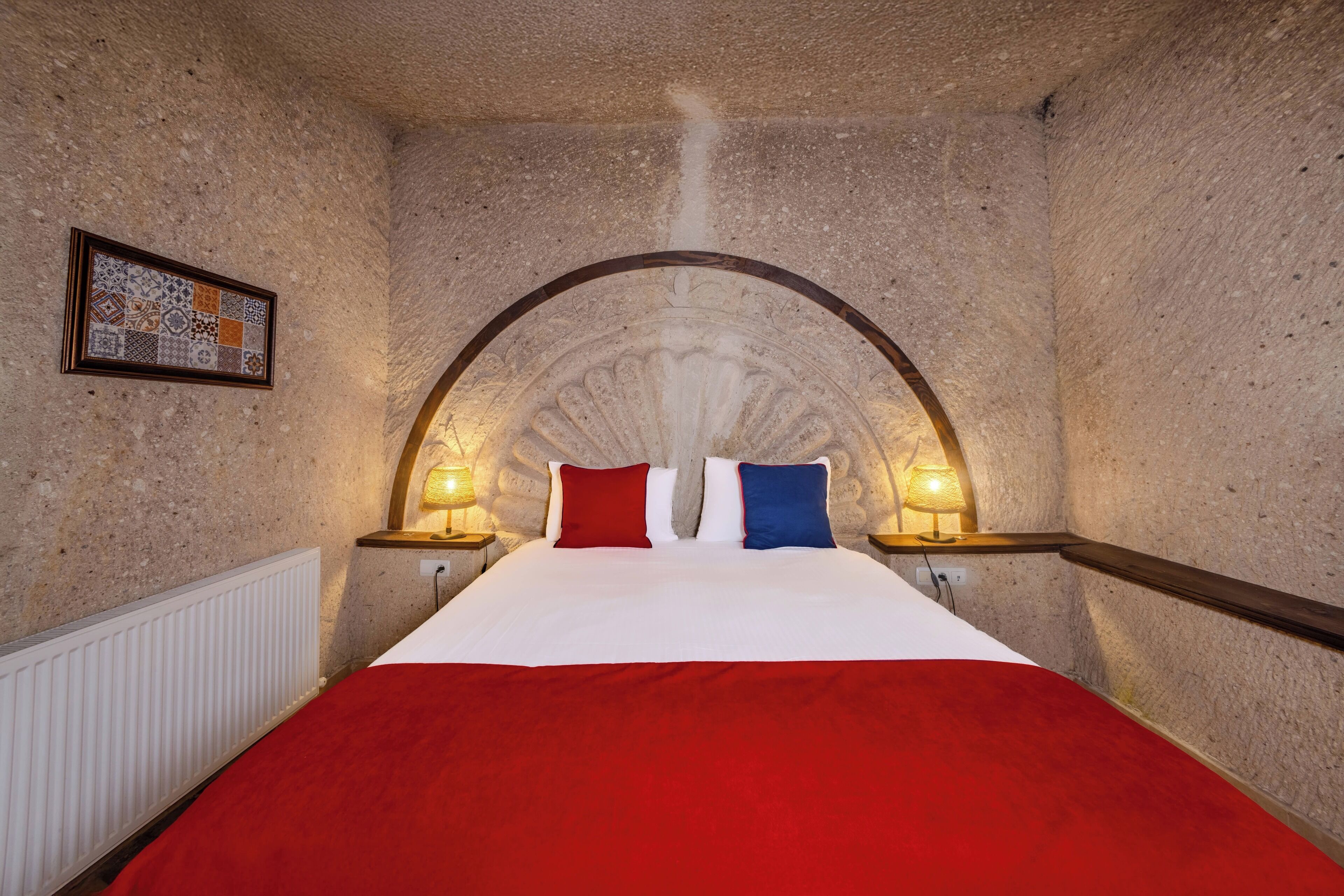 A room with a stone wall, a bed with a red and white bedspread, two bedside lamps, and a framed artwork on the wall.
