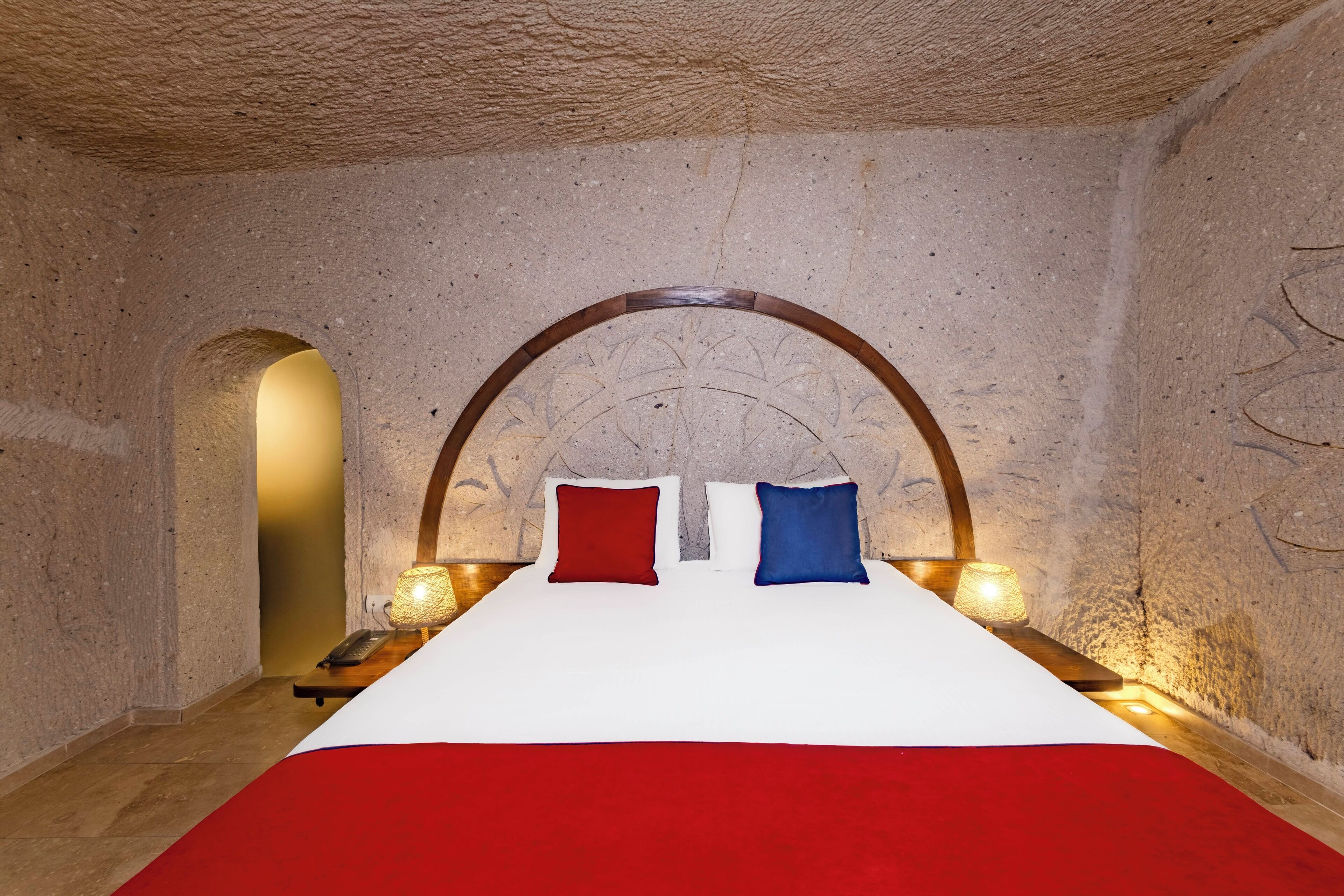A room with a stone wall, a bed with a white and red bedspread, two pillows, and two bedside lamps.