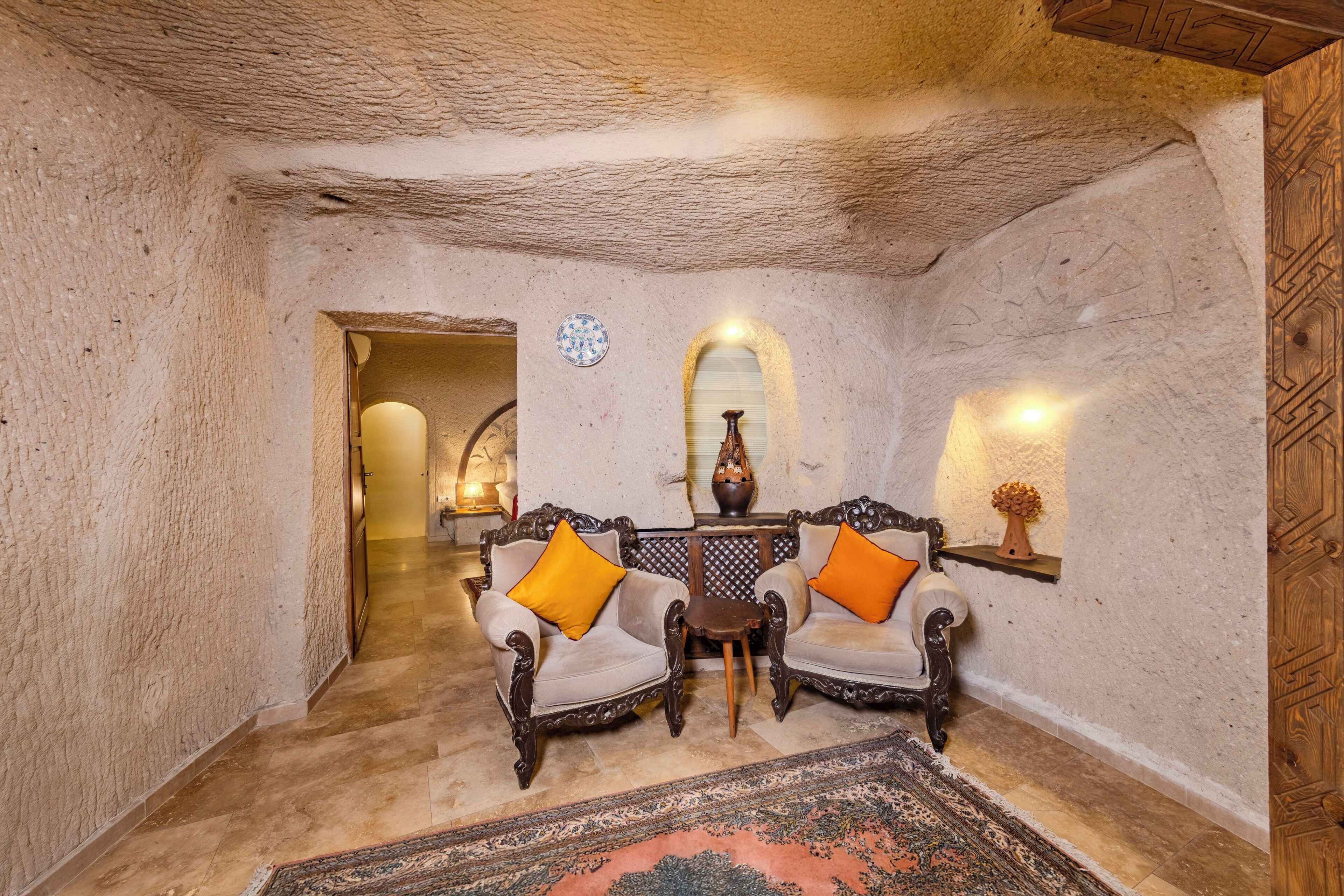 A room with rustic stone walls, a wooden ceiling, and a traditional rug. There are two ornate chairs with cushions, a small table, and a wall-mounted shelf with a vase.