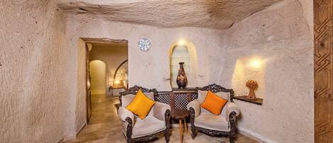 A room with rustic stone walls, a wooden ceiling, and a traditional rug. There are two ornate chairs with cushions, a small table, and a wall-mounted shelf with a vase.