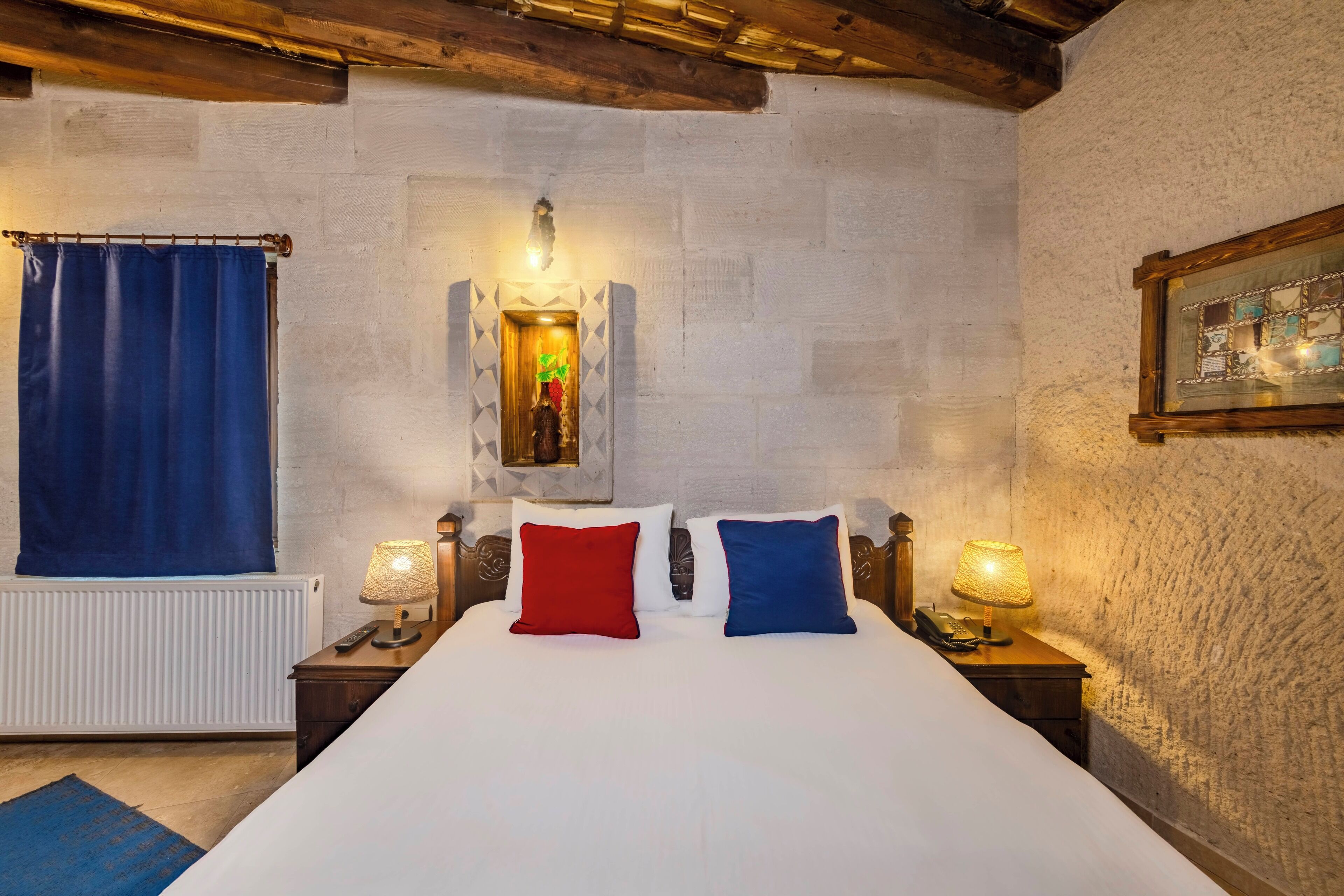 Una camera da letto con un letto grande, due comodini con lampade, una finestra con tenda blu, un termosifone e un muro in pietra.