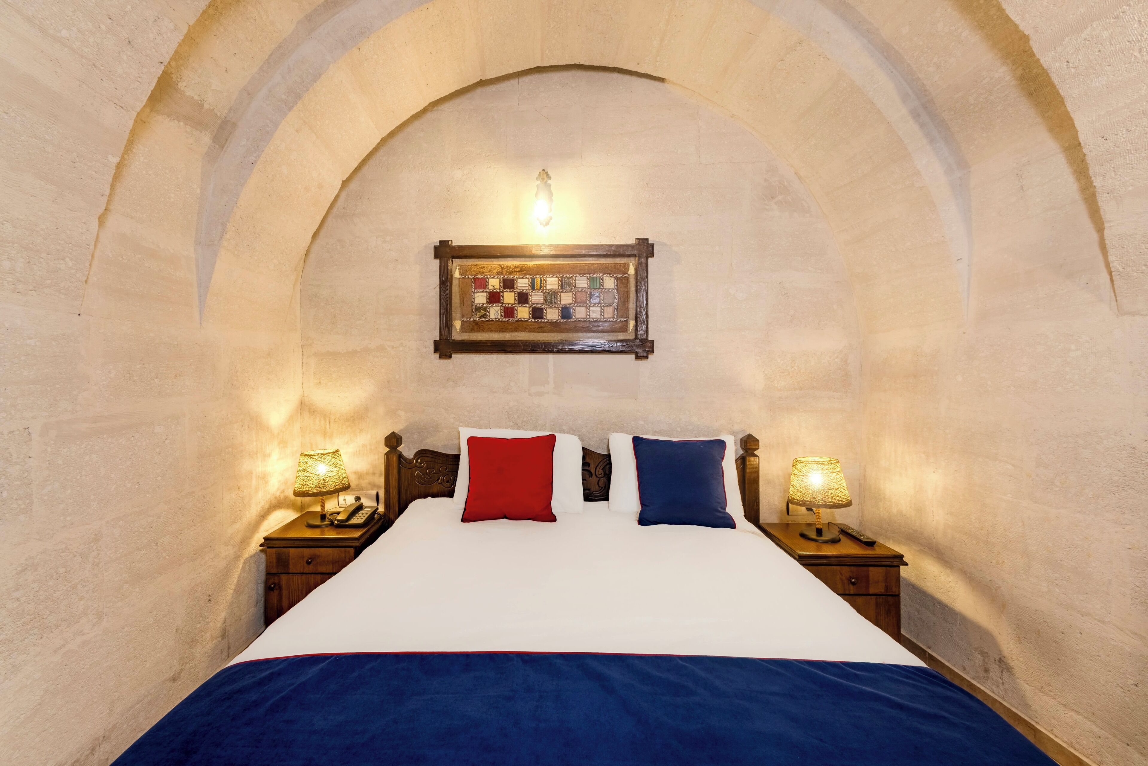 Un dormitorio con un arco de piedra, una cama de madera con colcha blanca y azul, dos lámparas de noche y un tablero de ajedrez enmarcado en la pared.