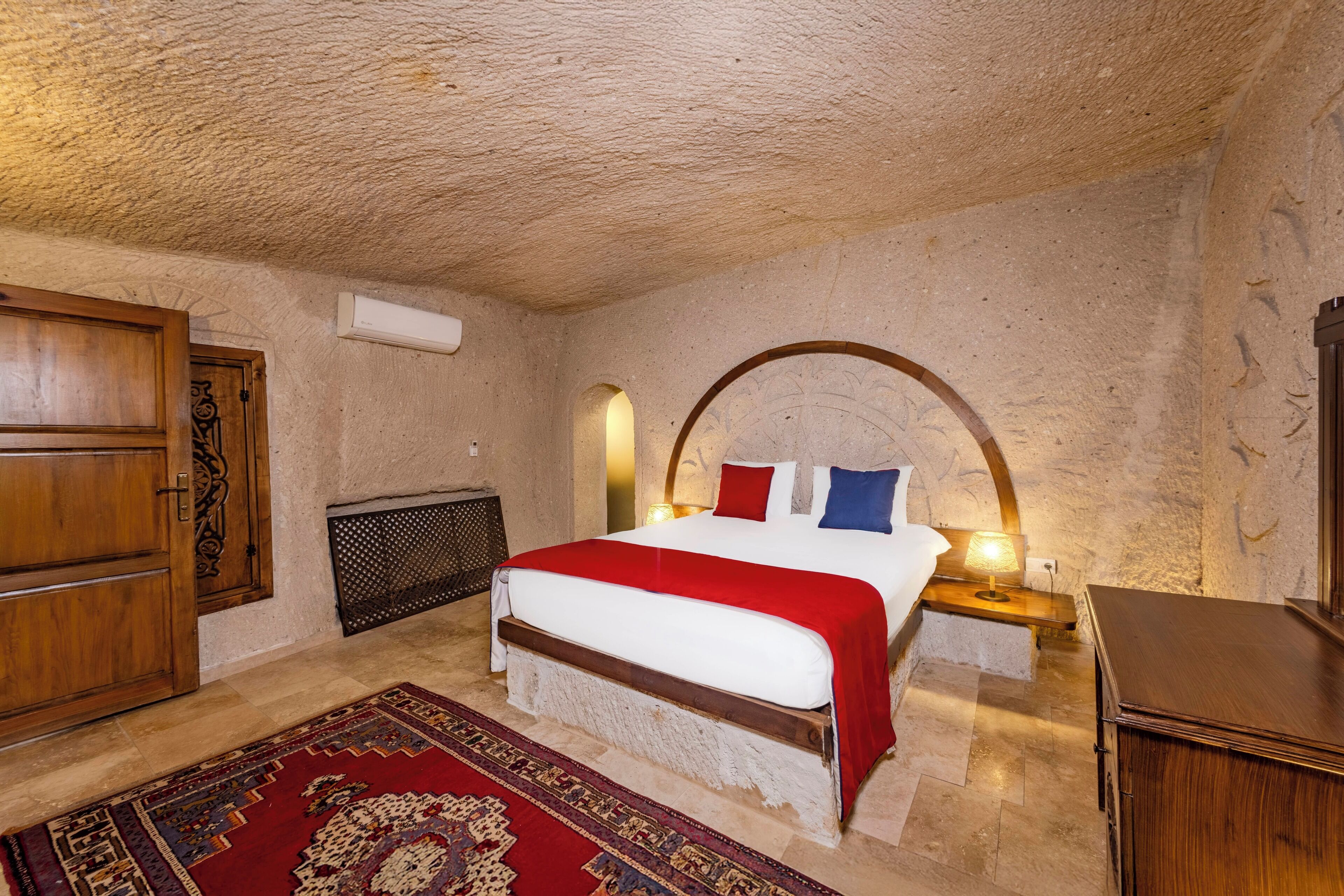 A bedroom with a large bed, a wooden wardrobe, a red and white bedspread, a patterned rug, and a stone wall.