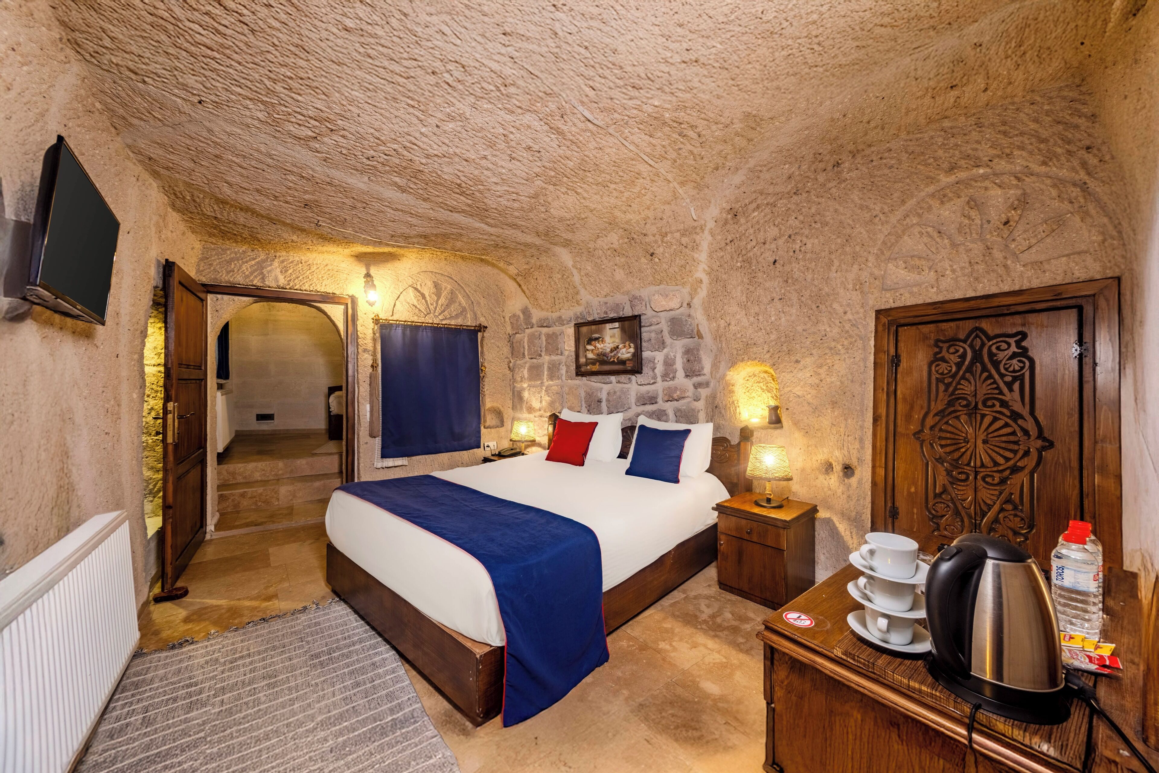 A bedroom with a stone ceiling, a bed with a blue blanket, a wooden nightstand with a kettle, and a door with an ornate design.