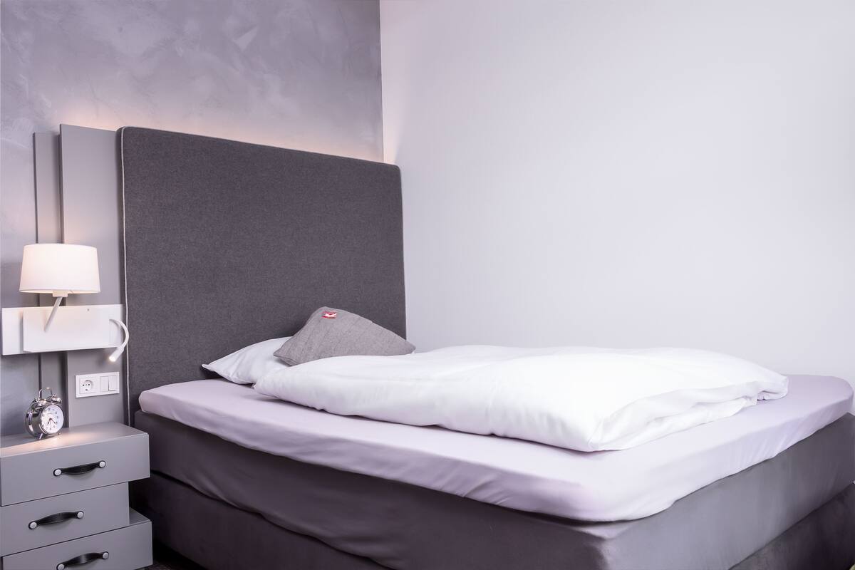 Una cama bien hecha con ropa de cama blanca, un cabecero gris y una lámpara de noche.