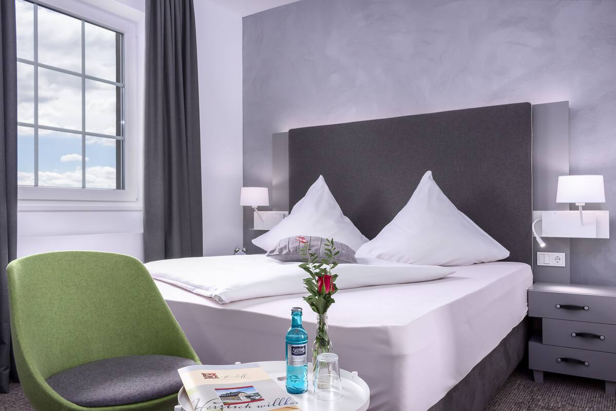 Habitación de hotel con una cama, una silla verde, una mesita de noche con una botella, un vaso y un jarrón con flores.