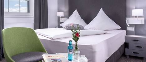 Habitación de hotel con una cama, una silla verde, una mesita de noche con una botella, un vaso y un jarrón con flores.