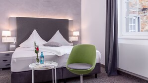 Une chambre d’hôtel avec un lit, deux lampes de chevet, un fauteuil vert, une petite table sur laquelle est posé un vase de fleurs et une fenêtre avec des rideaux.