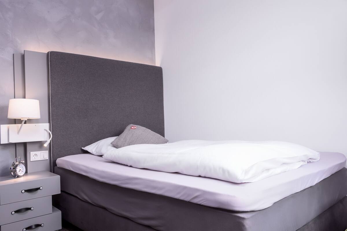 Una cama bien tendida con ropa blanca, un cabecero gris y una mesita de noche con una lámpara y un pequeño reloj.