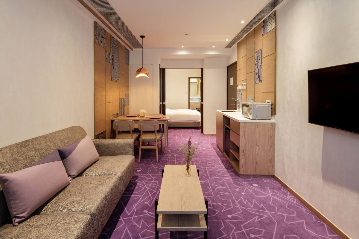A compact hotel room with a sofa, a small dining table, a bed, a TV, and a kitchenette.