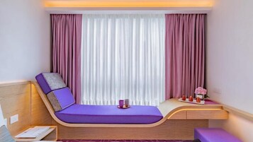 Una habitación moderna con una cama curvada, una mesita con un florero y una ventana con cortinas translúcidas.
