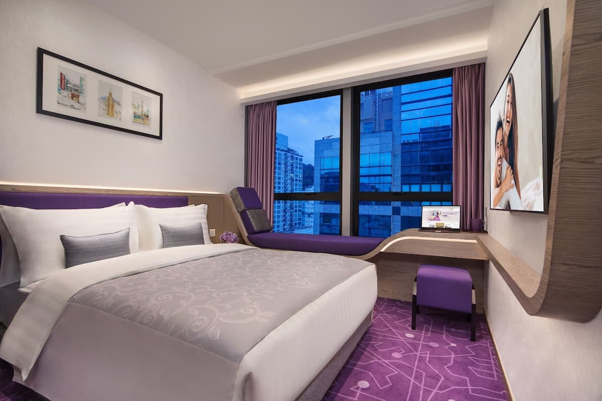 A modern hotel room with a large bed, a desk, and a view of city buildings through the window.
