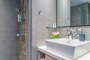 Un baño moderno con ducha, un lavamanos blanco y un estante de vidrio.