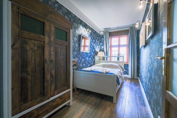 Une chambre avec une armoire en bois, un lit recouvert d’une courtepointe bleue et une fenêtre avec des rideaux.