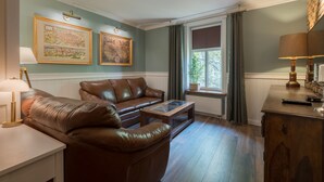 A living room with a brown leather sofa, a wooden coffee table, and a lamp on a side table. There are two framed artworks on the wall and a window with curtains. - Old House Apartments Poznań (Poznan)