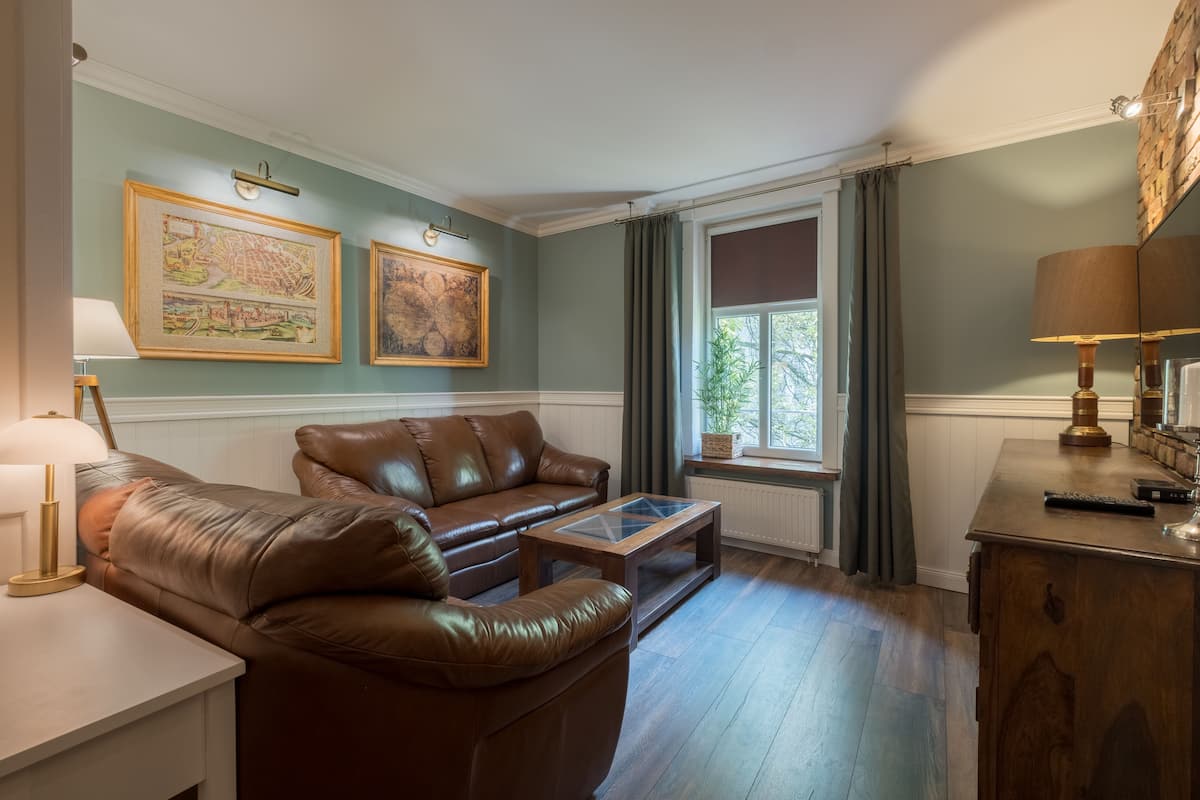 A living room with a brown leather sofa, a wooden coffee table, and a lamp on a side table. There are two framed artworks on the wall and a window with curtains.