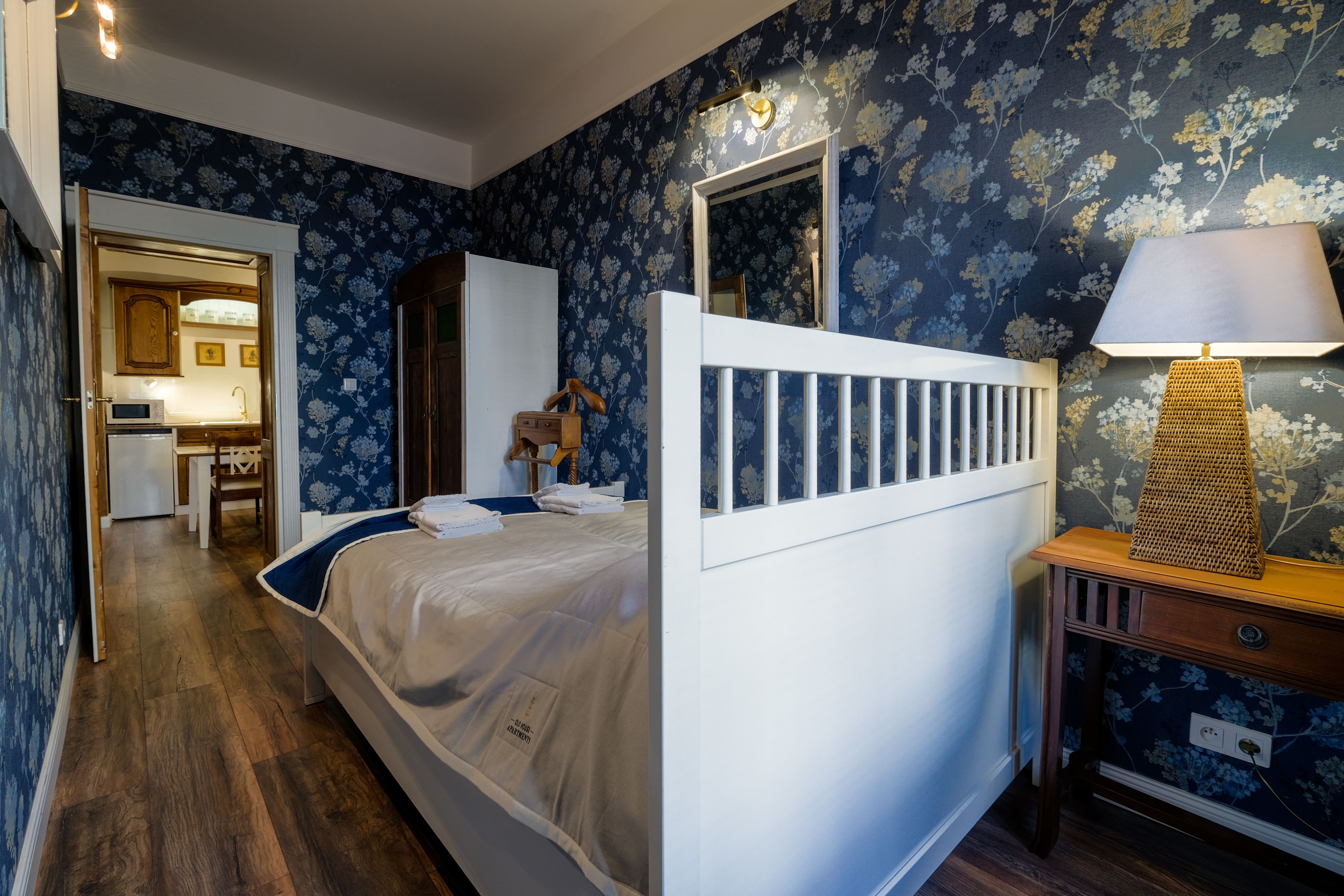 A bedroom with a bed, a bedside table with a lamp, a dresser, and a mirror. There is a door leading to a bathroom with a sink and a chair.