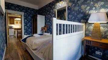 A bedroom with a bed, a bedside table with a lamp, a dresser, and a mirror. There is a door leading to a bathroom with a sink and a chair.