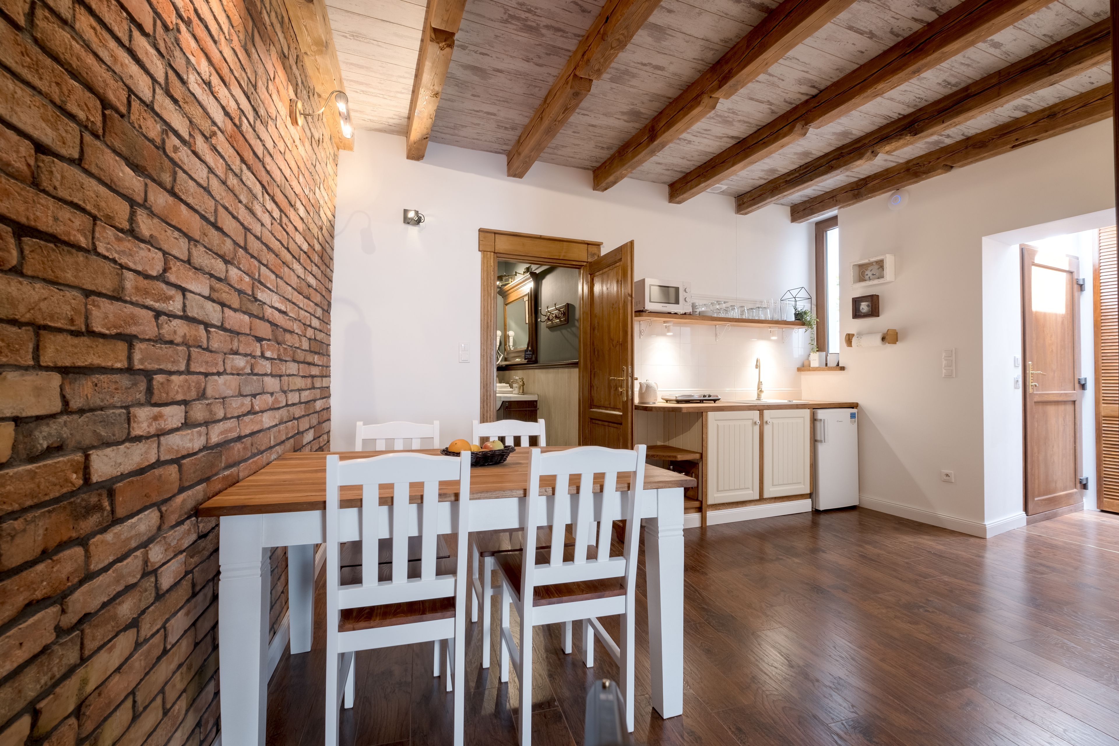 A dining area with a brick wall, wooden ceiling beams, and a wooden table with white chairs.