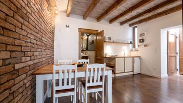 A dining area with a brick wall, wooden ceiling beams, and a wooden table with white chairs.