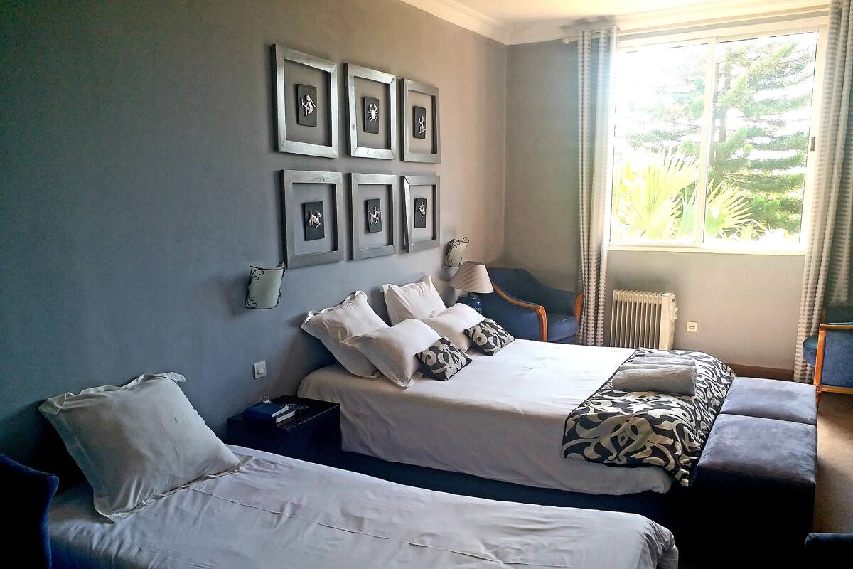 Une chambre d’hôtel avec deux lits, une fenêtre donnant sur un paysage verdoyant et des œuvres d’art encadrées accrochées au mur.