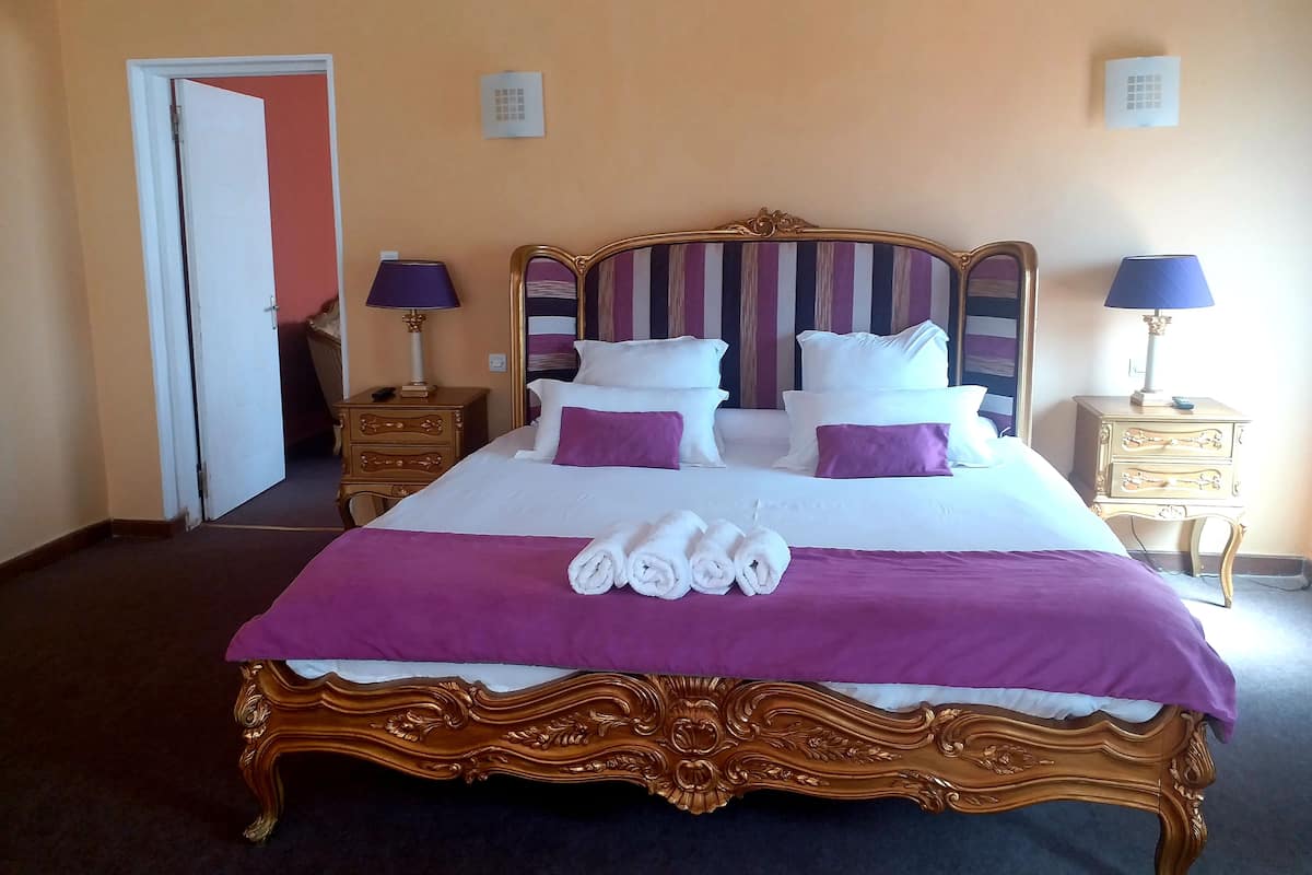 Un lit recouvert d’une couvre-lit violette et blanche, deux tables de chevet avec des lampes, et une porte visible au fond.