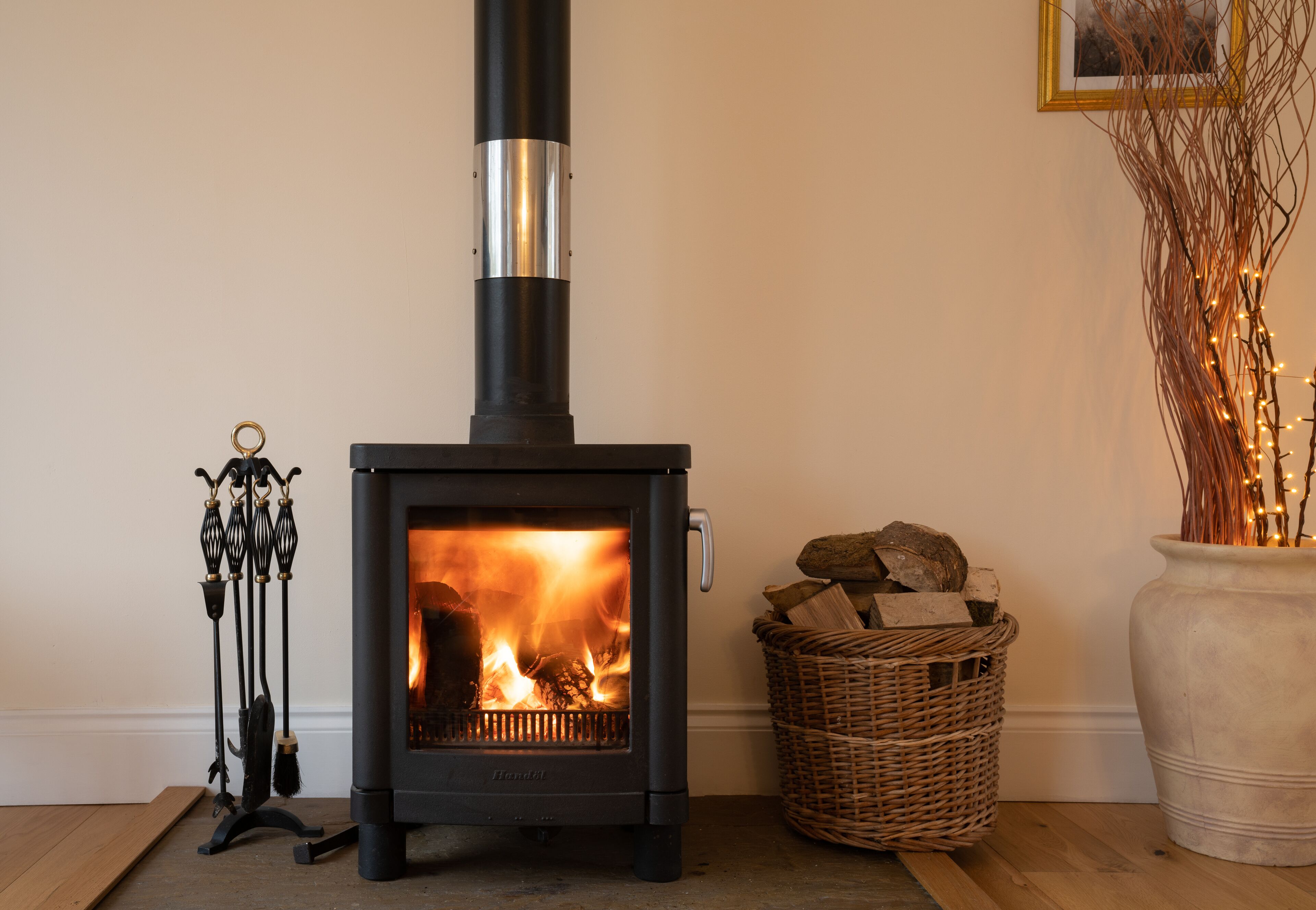 A wood-burning stove with a metal pipe, a wicker basket with logs, and a vase with branches.