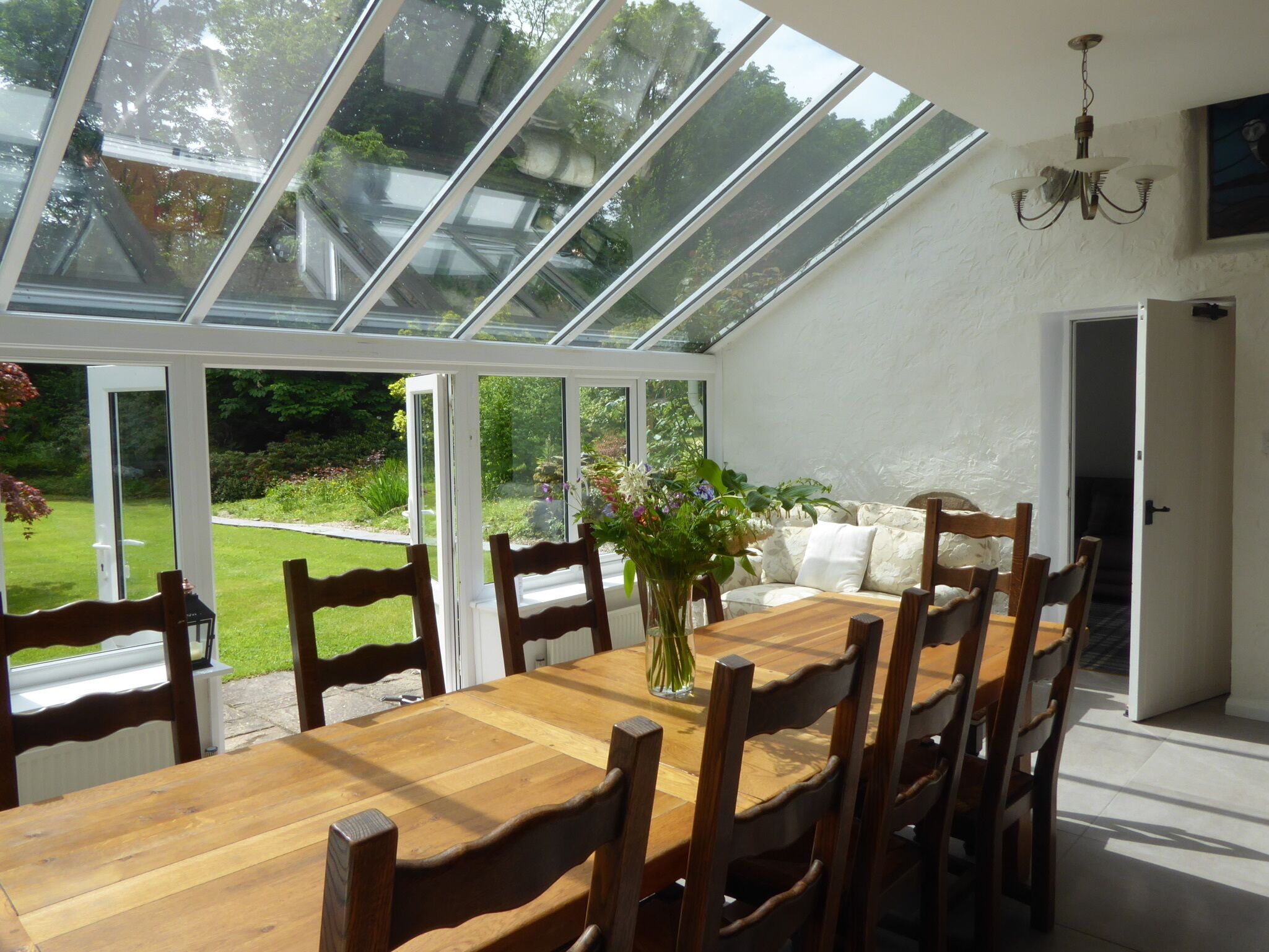 A conservatory with a wooden table and chairs, surrounded by white walls and a garden view.
