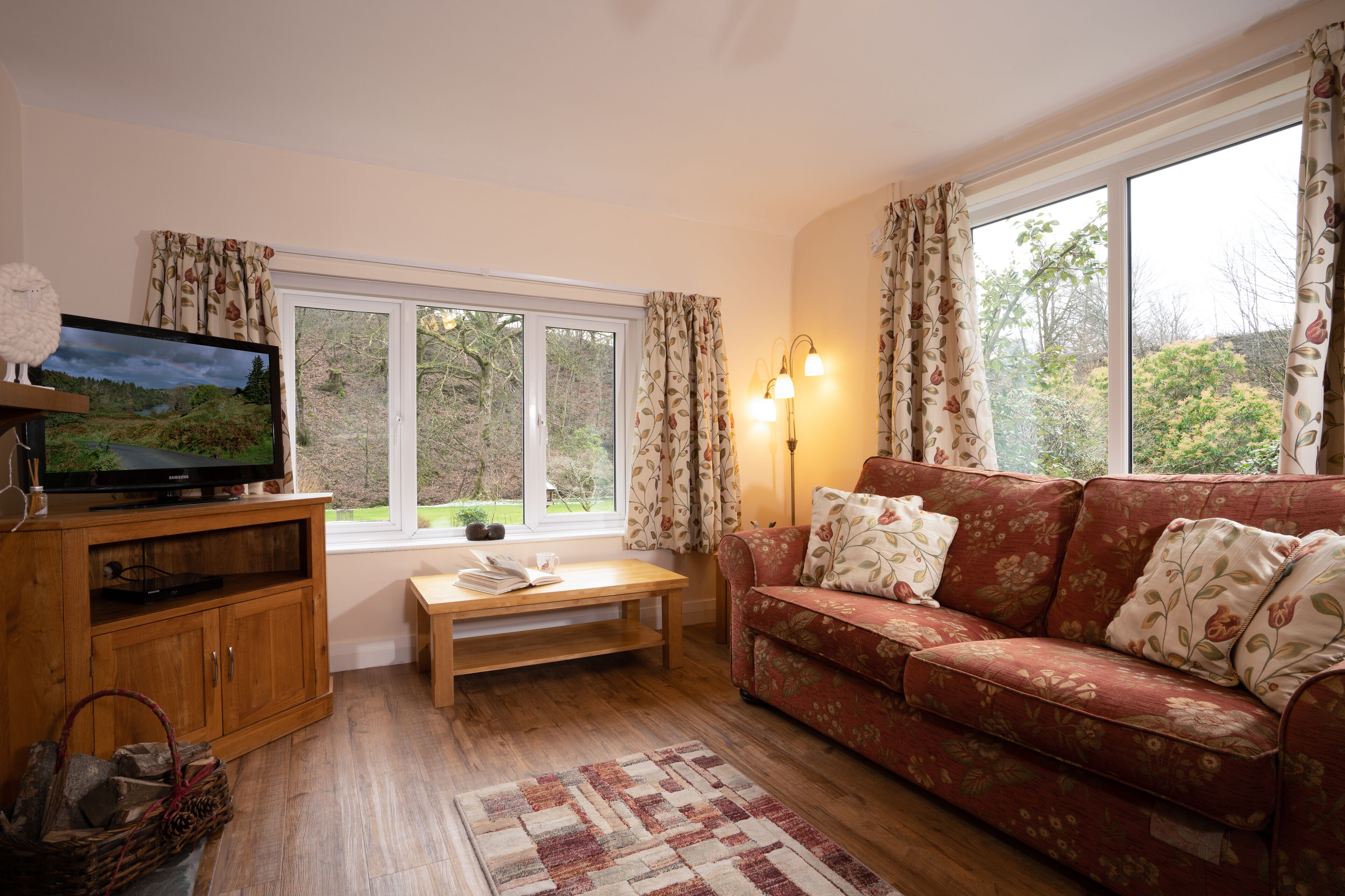 A cozy living room with a wooden TV stand, a red floral sofa, a wooden coffee table, and a view of greenery through large windows.