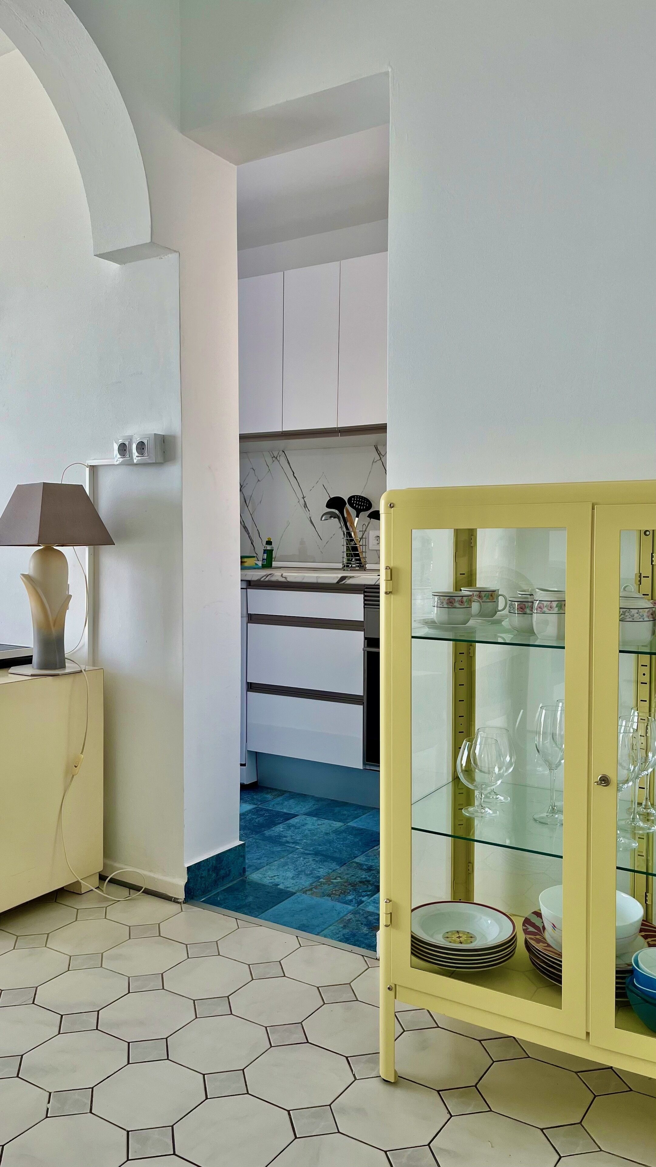 A modern kitchen with white cabinets, a blue-tiled floor, and a yellow cabinet with glass doors displaying dishes.